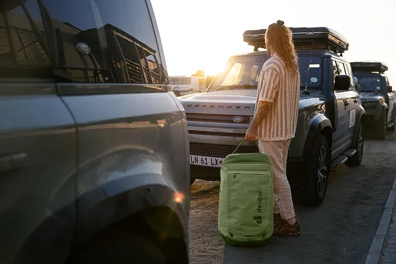 Junge Frau mit Reisegepäck vor einer Gruppe Geländewagen im Sonnenuntergang
